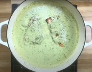 Salmon fillets cooking in creamy basil sauce in a white pot on a stovetop.